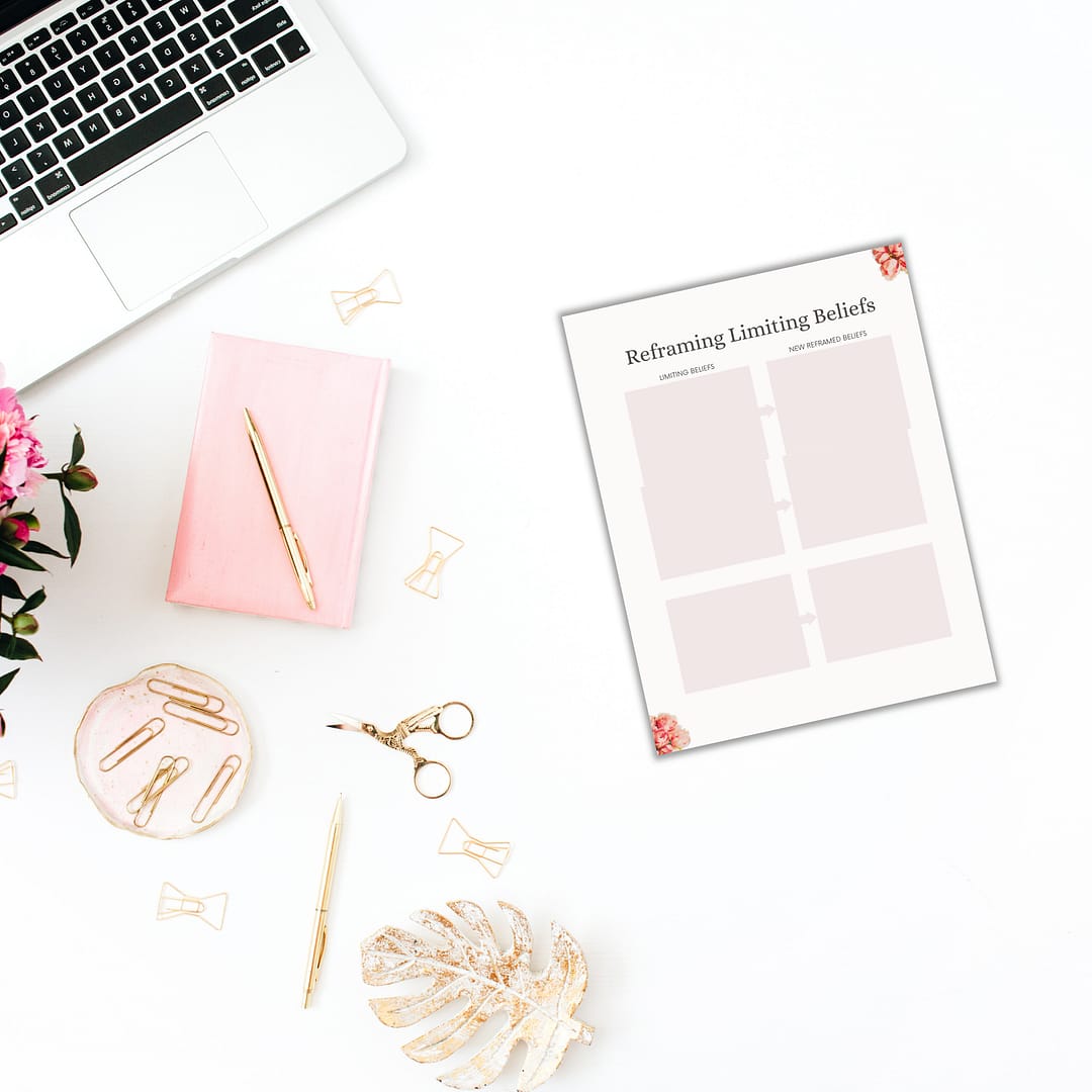 A workspace creatively arranged from a top-down perspective showcases a laptop, a pink notebook, a pen, paper clips, and scissors. Also visible is a sheet titled "Reframing Limiting Beliefs." Positioned nearby is the Vision Board Bundle filled with inspiring images. The setting is further enhanced by the presence of a pink flower and decorative leaf adding charm to the composition.