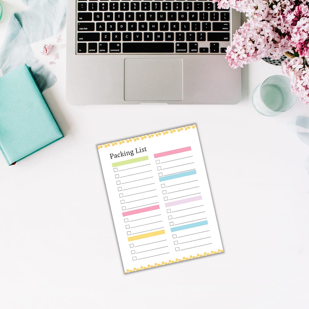 A laptop, a green notebook, and a packing list are arranged on a white surface with a bouquet of pink flowers nearby, capturing the essence of **Free: Spring-themed Planner Templates**.