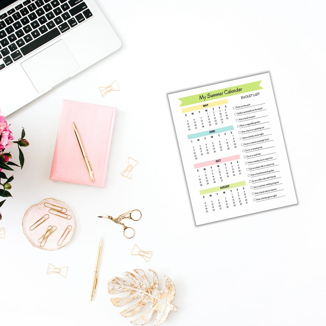 A neatly organized desk featuring a calendar, gold scissors, paper clips, a pink notepad, and a laptop makes the perfect setup for planning. Tucked under the pink flower in the corner is the "Summer Planner for Kids," ready to inspire fun activities for warm days ahead.