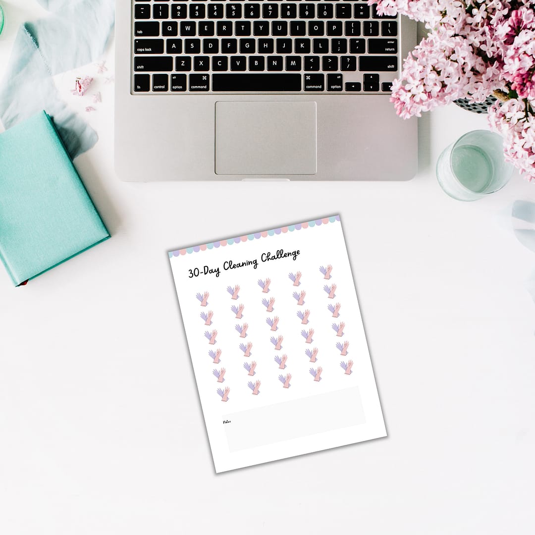 On a charming desk, an open laptop displays "The Cottagecore Cleaning Planner," a 30-day checklist that is perfectly complemented by a turquoise book and a vase of lilac flowers. This quaint setup embodies cottagecore vibes, combining rustic charm with the practical elegance of orderly living.