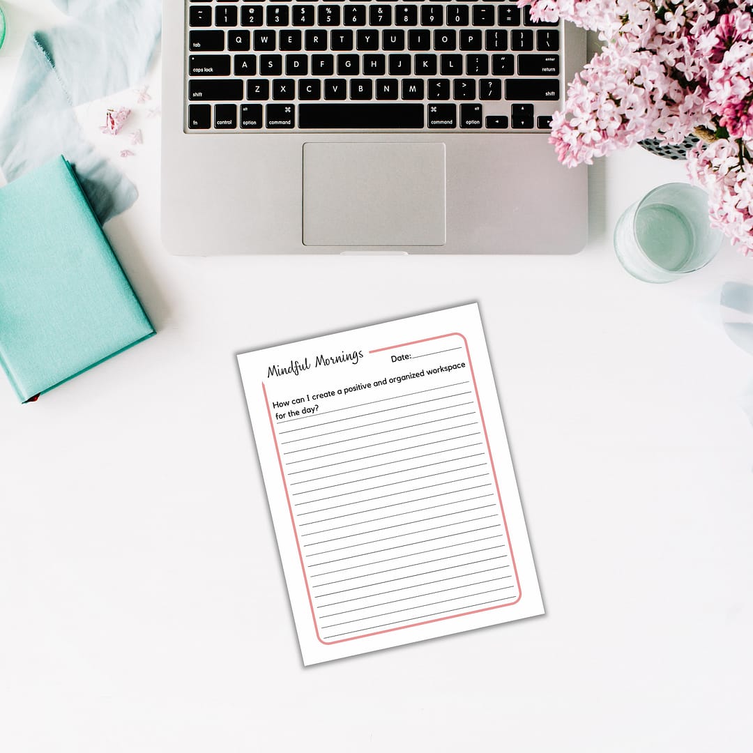A laptop sits on a white desk adorned with pink flowers and a turquoise journal. Nearby, the "Mindful Mornings Journal & Workbook" inspires thoughts on creating a positive workspace.