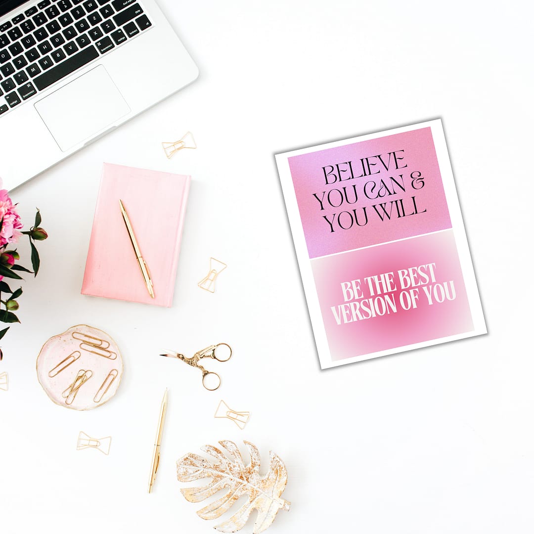 A flat lay of a desk showcases the Vision Board Bundle, including a laptop, pink notebook, and gold clips arranged elegantly with scissors. A leaf decoration enhances the display alongside pink flowers and a framed motivational quote, creating an inspiring setup to boost creativity.