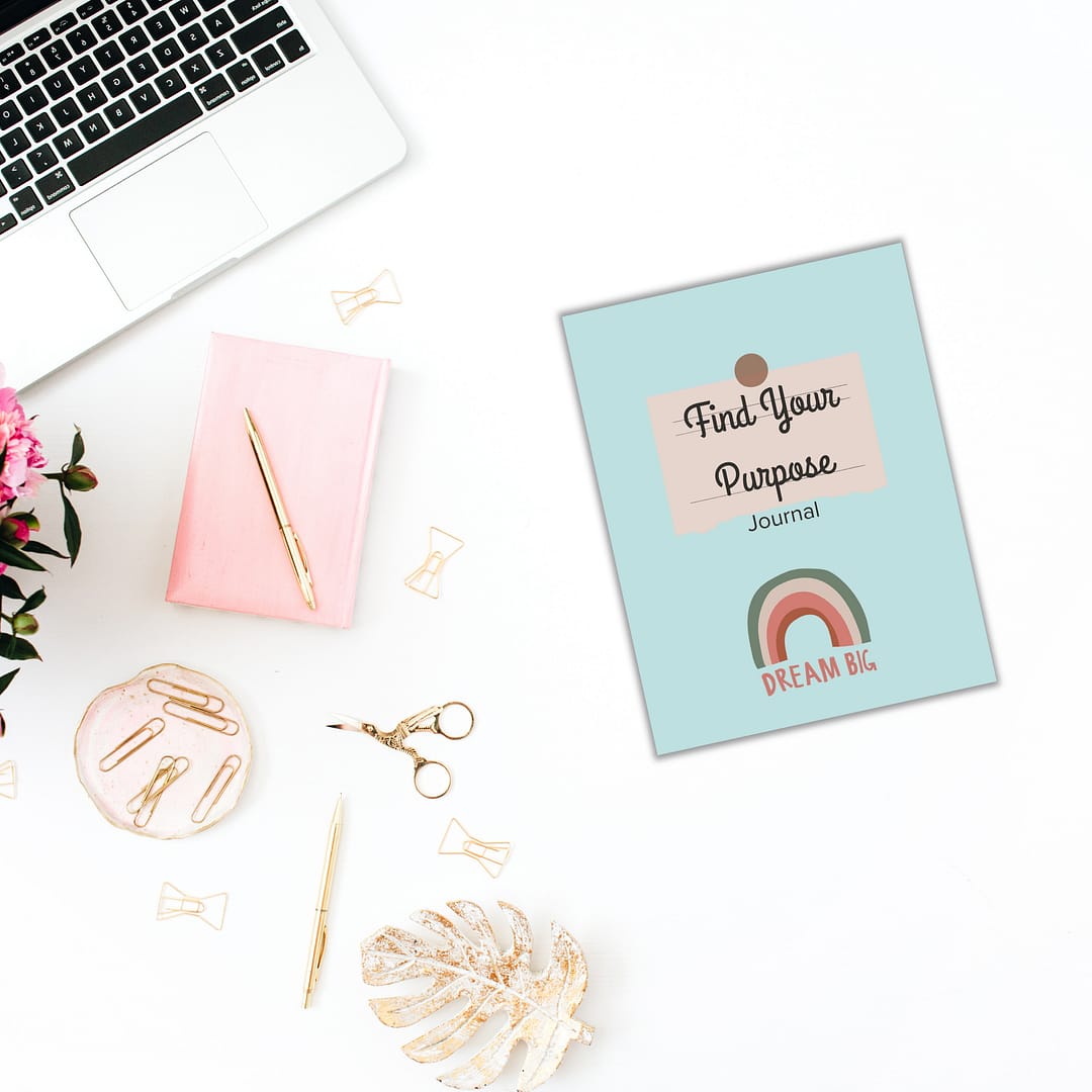 A desktop featuring a laptop, a pink notebook, and The 'Find Your Purpose' Journal is thoughtfully arranged amidst pens, paper clips, scissors, and a vibrant plant.