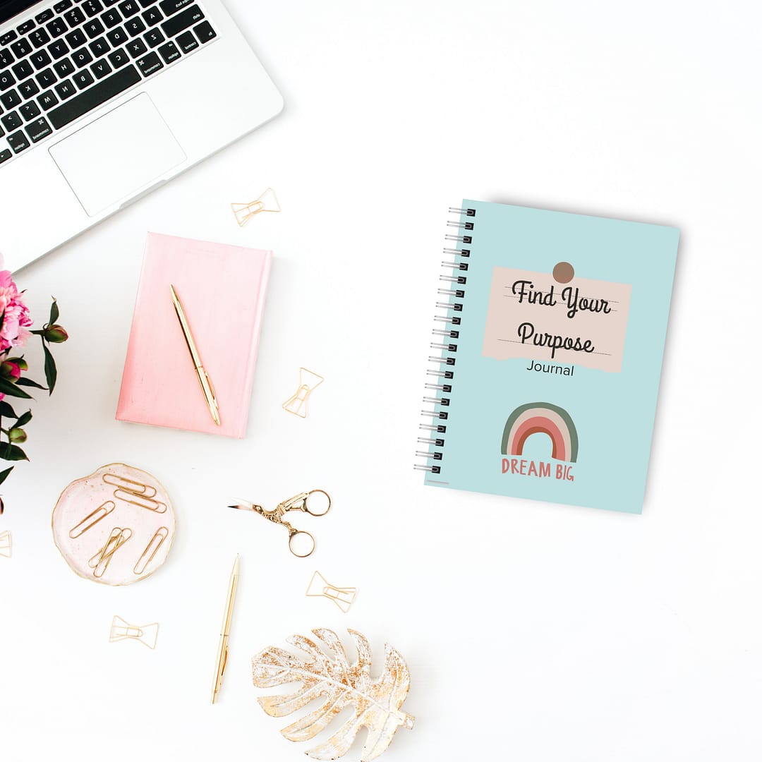 A laptop, pink notebook, and The 'Find Your Purpose' Journal are arranged on a white desk decorated with pink flowers, gold paperclips, and scissors.